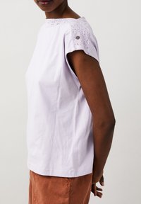 Fialový bavlněný top s krátkými rukávy, krajkovými detaily na ramenou a malým knoflíkovým akcentem. Texturovaná tkanina s uvolněným střihem.