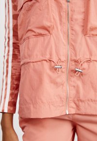 Ελαφρύ, ροζ κοραλί μπουφάν με κλείσιμο φερμουάρ, δύο μπροστινές τσέπες και κορδόνι στη μέση. Διαθέτει λευκές ρίγες στα πλάγια.