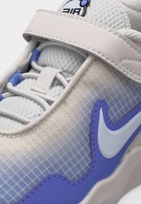 Λευκό και ανοιχτό μωβ αθλητικό παπούτσι με επάνω μέρος από δίχτυ, ρυθμιζόμενο λουράκι, λογότυπο Nike Swoosh και σήμανση «Air» στη γλώσσα.