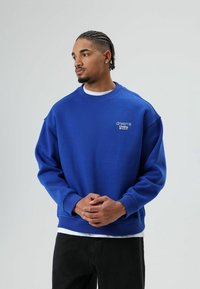 Sweat-shirt bleu à col rond en tissu doux, avec un logo blanc sur la poitrine, des poignets côtelés et une coupe ample. Porté sur une chemise blanche.
