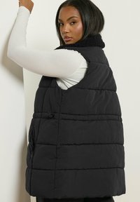 Svart pufferväst med en texturerad, fleecefoder i kragen, horisontellt kviltat mönster, sidofickor och en avslappnad passform.