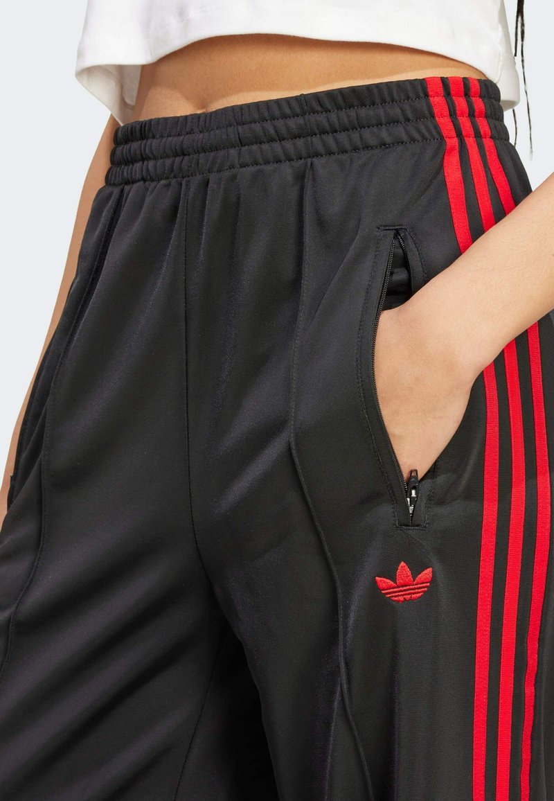 Pantalones deportivos negros con franjas laterales rojas, cinturilla elástica y bolsillos con cremallera. Presenta un pequeño logo rojo en la cadera izquierda.