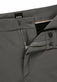 Un pantalon gris présente une fermeture éclair, un bouton de fermeture et des passants de ceinture. L'intérieur affiche un tissu contrastant avec une texture fine et des détails de couture.