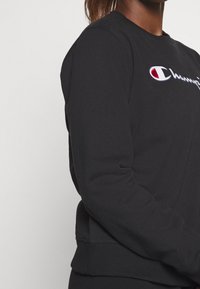 Sweat-shirt noir à manches longues, avec un logo Champion cousu en blanc et rouge, des poignets et un ourlet côtelés, et une texture douce.