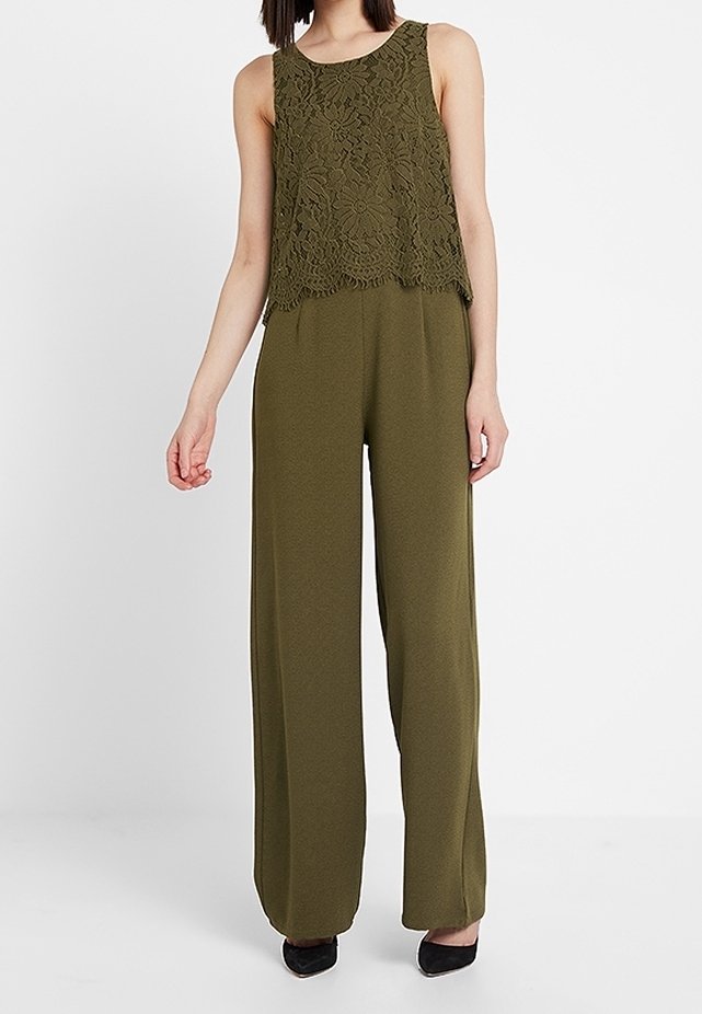 Femme portant un haut en dentelle sans manches vert olive avec un pantalon large assorti et des chaussures noires pointues sur fond blanc.