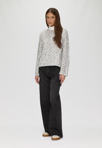 Pull en maille grise clair à la coupe décontractée, associé à un pantalon noir de style cargo. Le modèle se tient devant un fond uni.