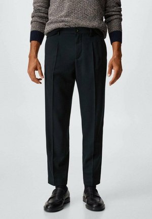 Uomo che indossa pantaloni neri su misura, mocassini neri e un maglione grigio con polsini blu navy, in piedi di fronte a uno sfondo chiaro e semplice.