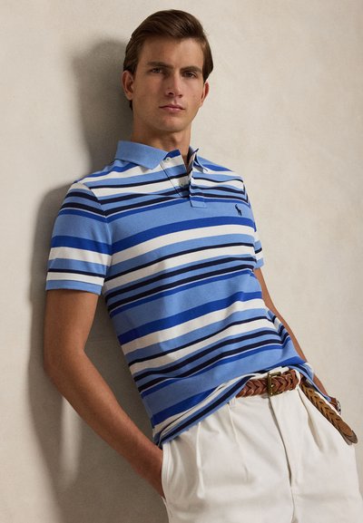 Homme appuyé contre un mur portant un polo rayé bleu, blanc et noir et un pantalon blanc avec une ceinture marron tressée, les mains dans les poches.