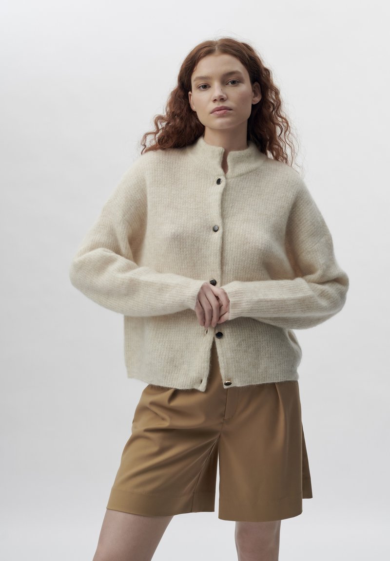 Cream-colored oversized cardigan made of soft wool, with a stand collar and front button closure. Paired with brown tailored shorts.