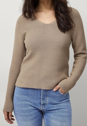 Femme portant un pull en maille beige à manches longues et un jean bleu clair, la main partiellement dans la poche, sur un fond clair uni.