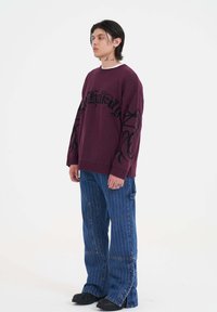 Bordový svetr s černým graffiti-style textem a vzory na rukávech, spárovaný s modrými pruhovanými kalhotami s širokými nohavicemi a černou obuví.