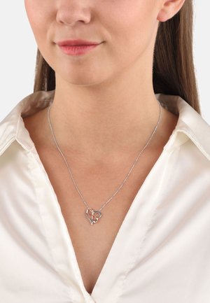 Collana d'argento a forma di cuore con design intrecciato su una delicata catena, indossata su una camicia bianca con colletto. Texture liscia, design moderno.