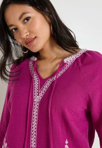 Tunique fuchsia avec un col en V, présentant des accents brodés blancs et des liens au niveau du col. Fabriquée en tissu texturé, elle a des manches longues.