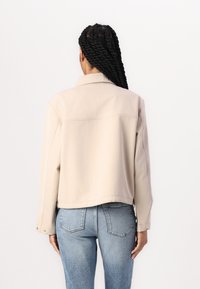 Beige zkrácená bunda s klasickým límcem, předními kapsami a knoflíkovými manžetami, v kombinaci se světle modrými džínami s vysokým pasem.