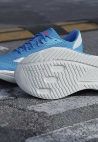 Chaussure de sport bleue avec semelle blanche montrant un profil texturé, posée sur l’asphalte avec un dos d’âne et des marquages routiers en arrière-plan.