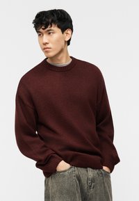 Jeune homme aux cheveux bouclés et foncés, portant un pull en laine bordeaux et un pantalon gris délavé, les mains dans les poches, regardant vers sa gauche.