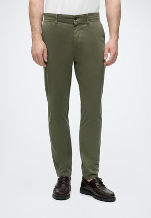 Férfi olivazöld slim-fit nadrágban és sötétbarna bőr fűzős cipőben, egyszínű háttér előtt áll.