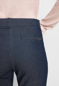 Gros plan d’une personne portant un pantalon bleu foncé sur mesure et un haut texturé rose clair à manches longues, montrant les détails de la poche arrière et de la ceinture.