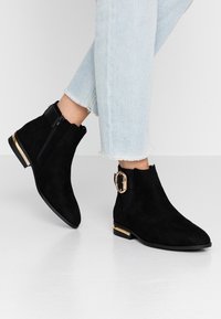 Botines de ante negro con cremallera lateral y hebilla con acentos dorados. Suela plana, punta afilada y tacón corto. Diseño casual.