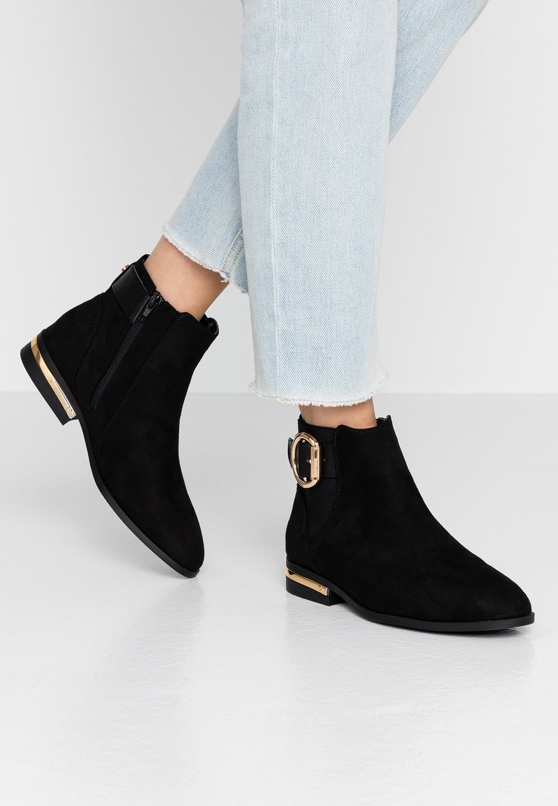 Botines de ante negro con cremallera lateral y hebilla con acentos dorados. Suela plana, punta afilada y tacón corto. Diseño casual.