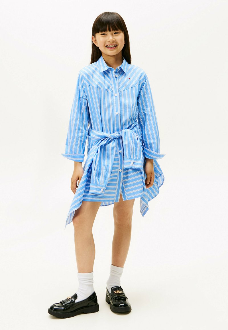Abito camicia a righe blu e bianche con maniche lunghe, chiusura a bottoni e cintura in vita. Abbinato a mocassini neri e calzini bianchi.