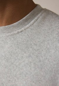 Hellgrauer Sweatshirt mit einem weichen, strukturierten Stoff. Verfügt über einen gerippten Rundhalsausschnitt und leichte Flecken im Material für zusätzliche Details.