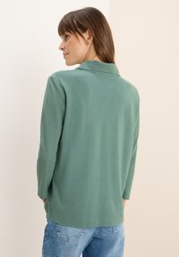 Camicia verde a maniche lunghe con colletto, realizzata in tessuto morbido. Presenta fessure laterali all'orlo e semplici dettagli di cucitura, abbinata a jeans blu.