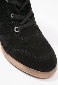 Gros plan d'une basket en daim noir avec une couture en zigzag, des lacets noirs, des œillets en métal et une semelle en caoutchouc texturée marron clair.