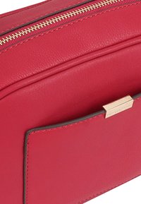 Rode leren tas met een gouden rits, gladde textuur en een voorvak met een metallic accent. Eenvoudig, rechthoekig ontwerp.