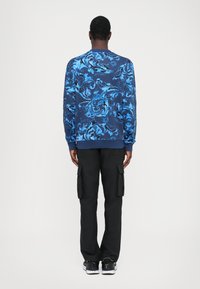 Langermet blå blomster genser med intrikate mønstre, sammen med svarte cargo bukser, sett fra ryggen. Fritids joggesko.