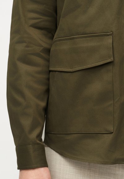 Κοντινό πλάνο μανσετών από μπουφάν σε ελαιοπράσινο χρώμα και μεγάλο τσέπη με καπάκι, φορεμένα με ανοιχτό μπεζ παντελόνι.