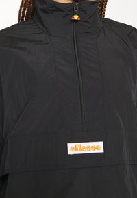 Czarny pulower z zamkiem ćwierć, wykonany z lekkiego, błyszczącego materiału. Posiada haftowane logo "ellesse" w kolorze pomarańczowym oraz kolorowy uchwyt suwaka.