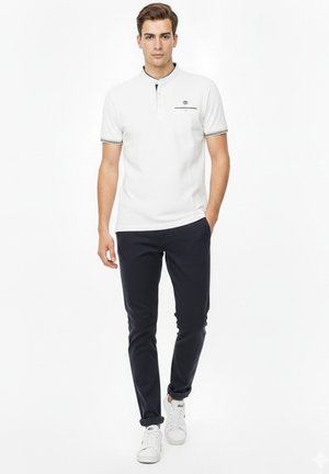 Joven con un polo blanco de manga corta, pantalones ajustados oscuros y zapatillas blancas, posando contra un fondo blanco.