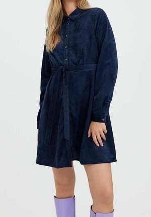 Femme portant une robe chemise en velours côtelé bleu marine avec ceinture à la taille et des bottes lavande montant jusqu'aux genoux, debout devant un fond uni.