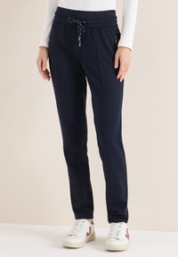 Marineblauwe joggingbroek met een taillekoord, twee voorzakken en geribbelde manchetten, gecombineerd met witte sneakers met rode accenten.