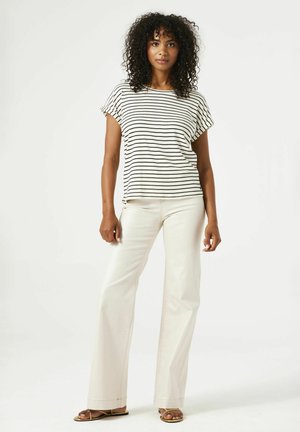 Top à manches courtes rayé en crème et noir, associé à un pantalon large de couleur claire. Cheveux bouclés naturels, en arrière-plan uni.