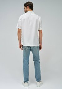 Camicia bianca a maniche corte in tessuto liscio, abbinata a jeans blu chiaro; la vista posteriore mostra una vestibilità comoda e dettagli di design minimi.