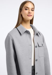 Grijze, wollen mixjacket met een kraag, mooie knopen aan de voorkant, een klepzak en textuurstof, gedragen over een lichtgekleurde top.