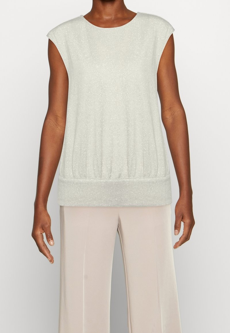 Haut sans manches gris clair avec un large ourlet porté avec un pantalon large beige clair sur une personne debout devant un fond uni.