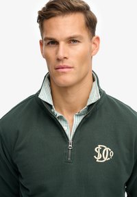 Pull-over vert foncé avec une fermeture éclair au quart, arborant un logo cousu sur le côté gauche de la poitrine et une chemise rayée à col en dessous.