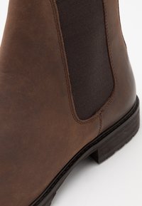 Ruske nahke čevlje do gležnja, ki imajo elastične stranske panele, teksturirano površino in trden gumijast podplat. Vidni šivi izboljšujejo dizajn.