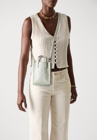 Helles grünes Kunstledertasche mit geprägtem Muster, ausgestattet mit kurzen Griffen und einem abnehmbaren Schultergurt, getragen über einem strukturierten cremfarbenen Pullover.