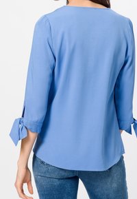 Blouse bleu clair avec des manches trois-quarts, présentant des détails à nouer aux poignets et un ourlet arrondi. Le tissu semble lisse et doux.
