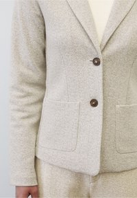 Saka v herringbone vzoru ve světle béžové barvě, se dvěma předními kapsami, zapínáním na jeden knoflík a přiléhavým střihem. Hladká textura.