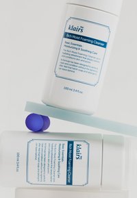 Bouteille en plastique blanche avec texte bleu, forme cylindrique, 100 ml. L'étiquette présente le nom du produit et les détails. Positionnée sur une surface bleu clair avec une sphère violette.