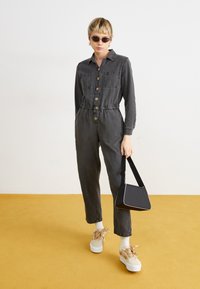Jumpsuit in denim grigio scuro con maniche lunghe, chiusura frontale a bottoni, vita elastica, tasche laterali, abbinato a sneaker platform beige e una borsa nera.