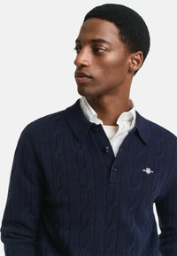 Jonge man met kort krullend haar in een marineblauwe kabeltrui over een witte blouse, die naar de zijkant kijkt.