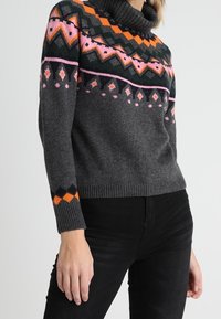 Šedý pletený svetr s tunelovým výstřihem. Obsahuje barevné geometrické vzory v oranžové, růžové a zelené barvě na hrudi a rukávech.