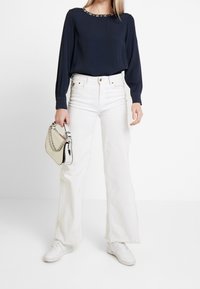 Eine Frau steht vor einem neutralen Hintergrund, trägt ein navyblaues Langarmoberteil, weiße Weitbeinhosen und hält eine kleine weiße Handtasche.
