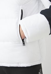 Hvid pufferjakke med en sort detalje, som har en lynlåslomme og et struktureret, polstret design på ærmerne og kroppen.